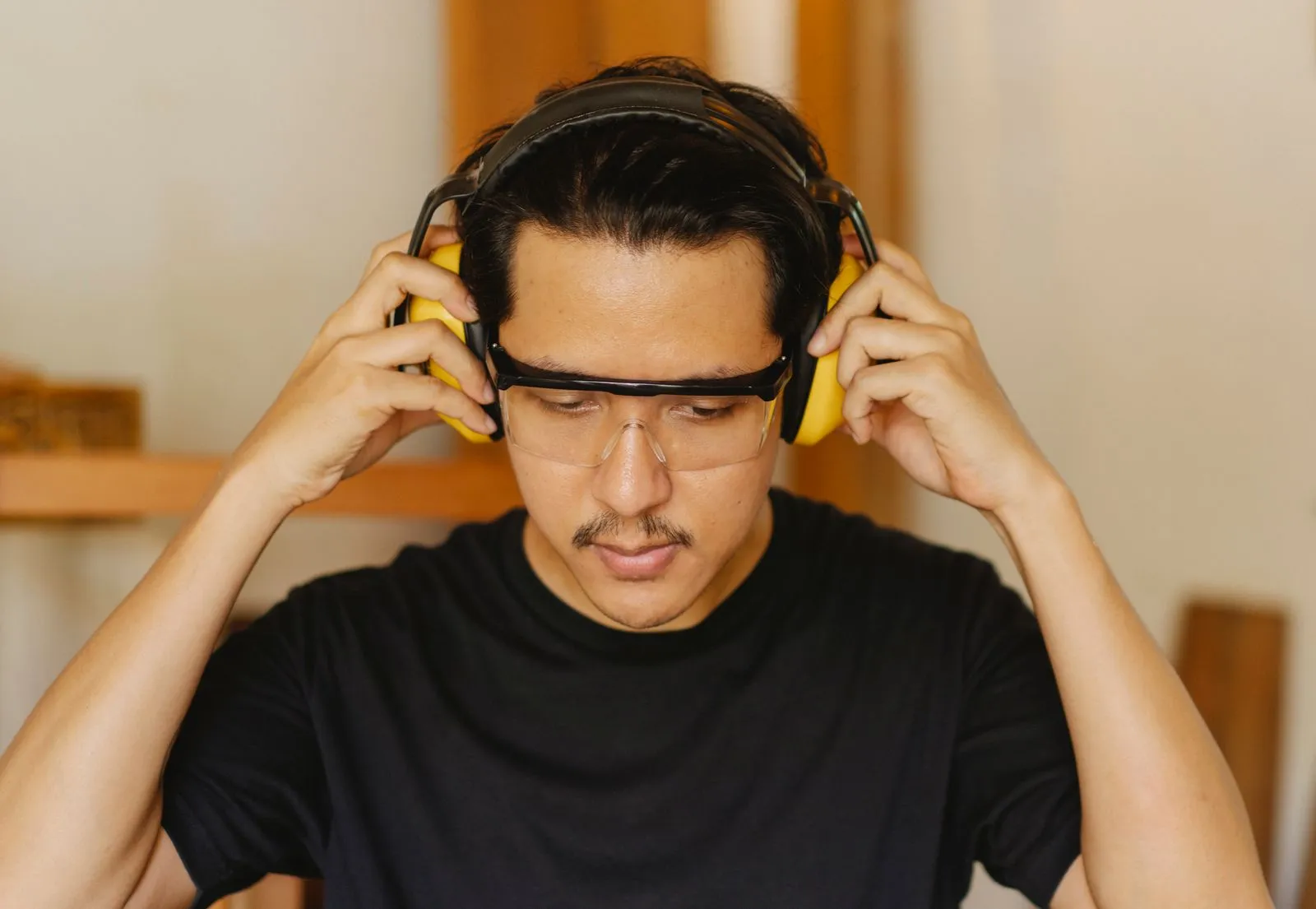 Young concentrated Asian male carpenter in protective glasses putting on headphones while looking down in workshop